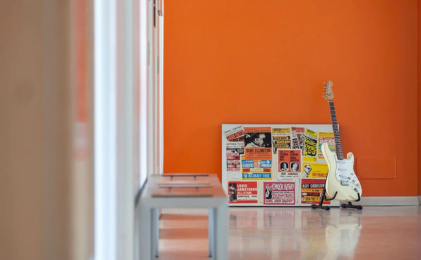 Chitarra elettrica bianca appoggiata a un muro arancione, accanto a una cornice con poster vintage di artisti storici, nel corridoio della Scuola e Accademia di Musica Cluster