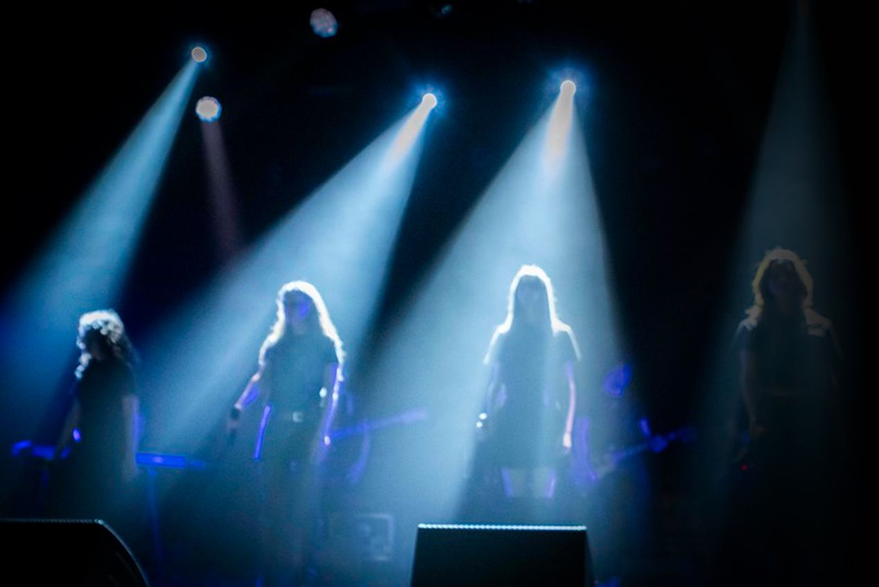 Allieve della Scuola e Accademia di Musica Cluster sul palco durante una performance dal vivo, illuminate da fasci di luce blu in un'atmosfera concertistica